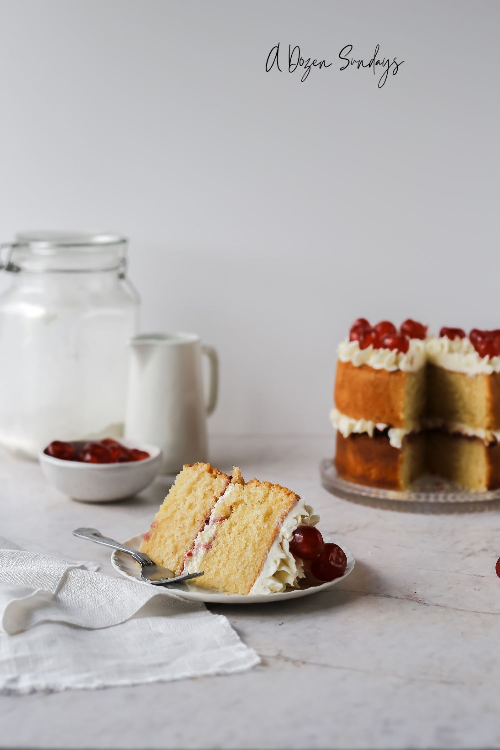 A slice of cake on a plate with the cake behind - easy Cherry Bakewell cake recipe from A Dozen Sundays