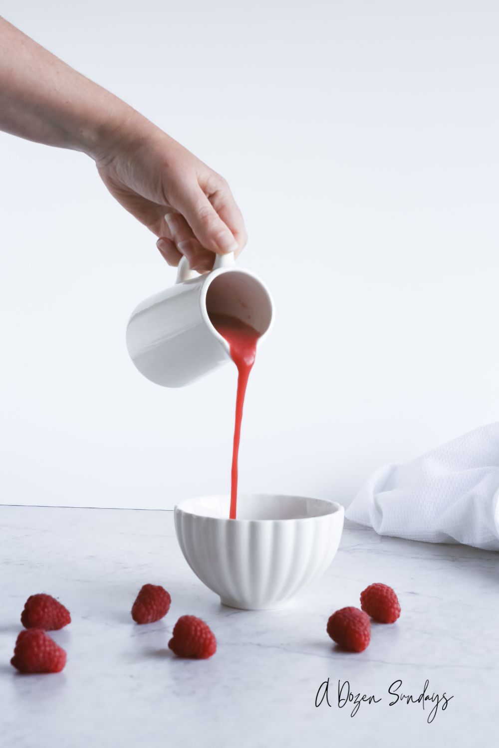 Raspberry Coulis being poured from a white jug into a white scallop edged bowl - Easy Homemade Raspberry Coulis Recipe