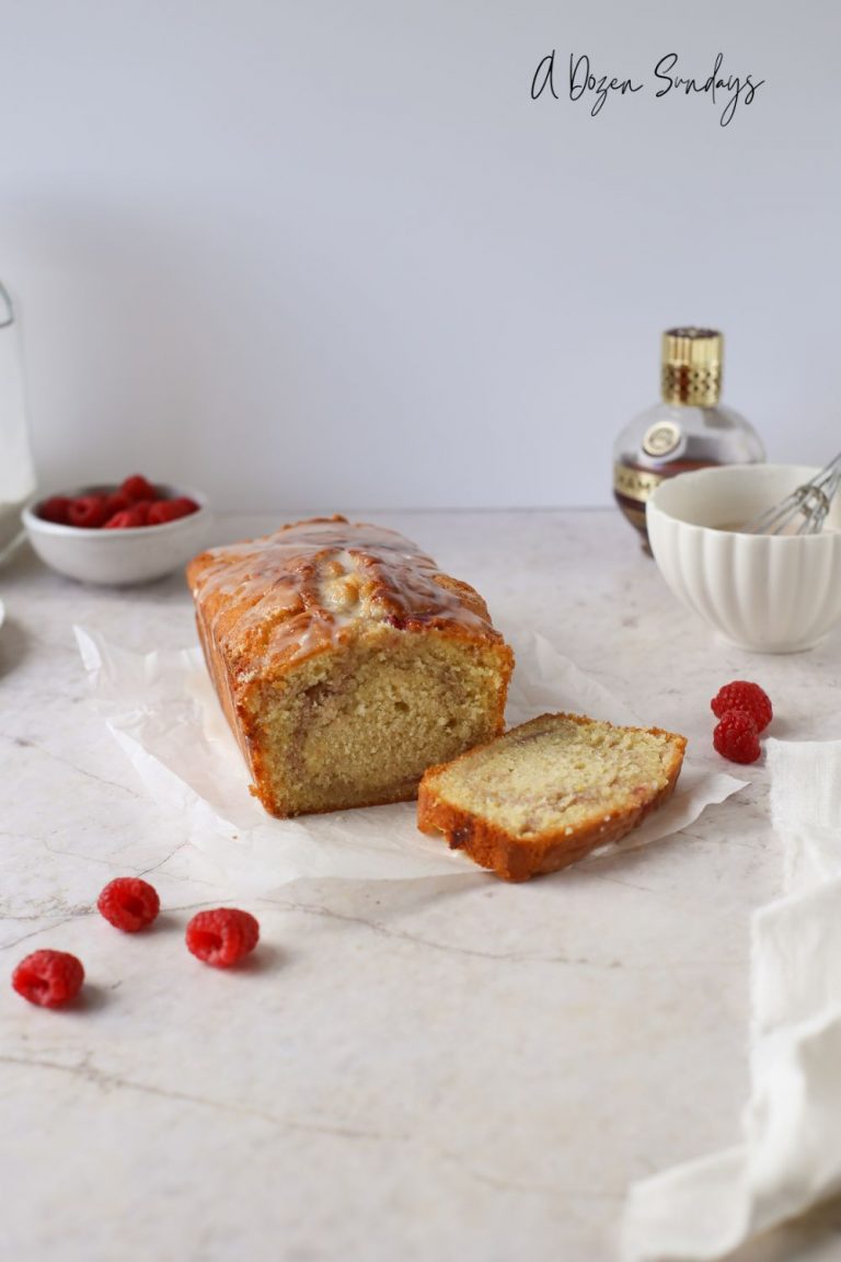 Lemon and Raspberry Loaf Cake with loose raspberries and baking equipment surrounding it - A Dozen Sundays